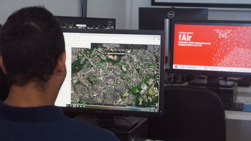 The image shows the back of a participant while he engages in testing fair in a desktop computer in front of him. His display shows a high resolution aerial image, while a screen in the backdrop has a sign that says f